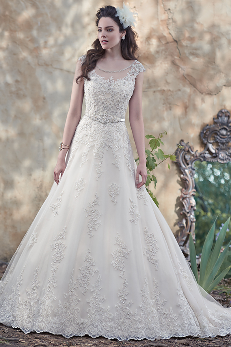 Glimmering lace appliqués, dotted with Swarovski crystal and pearl embellishments, lay atop tulle in this alluring ball gown, with stunning illusion sweetheart neckline and illusion back. Dainty lace cap-sleeves and a delicate ribbon belt at the waist complete the look. Finished with covered buttons over zipper and inner elastic closure.
<a href="https://www.maggiesottero.com/maggie-sottero/morgan/9512" target="_blank">Maggie Sottero</a>