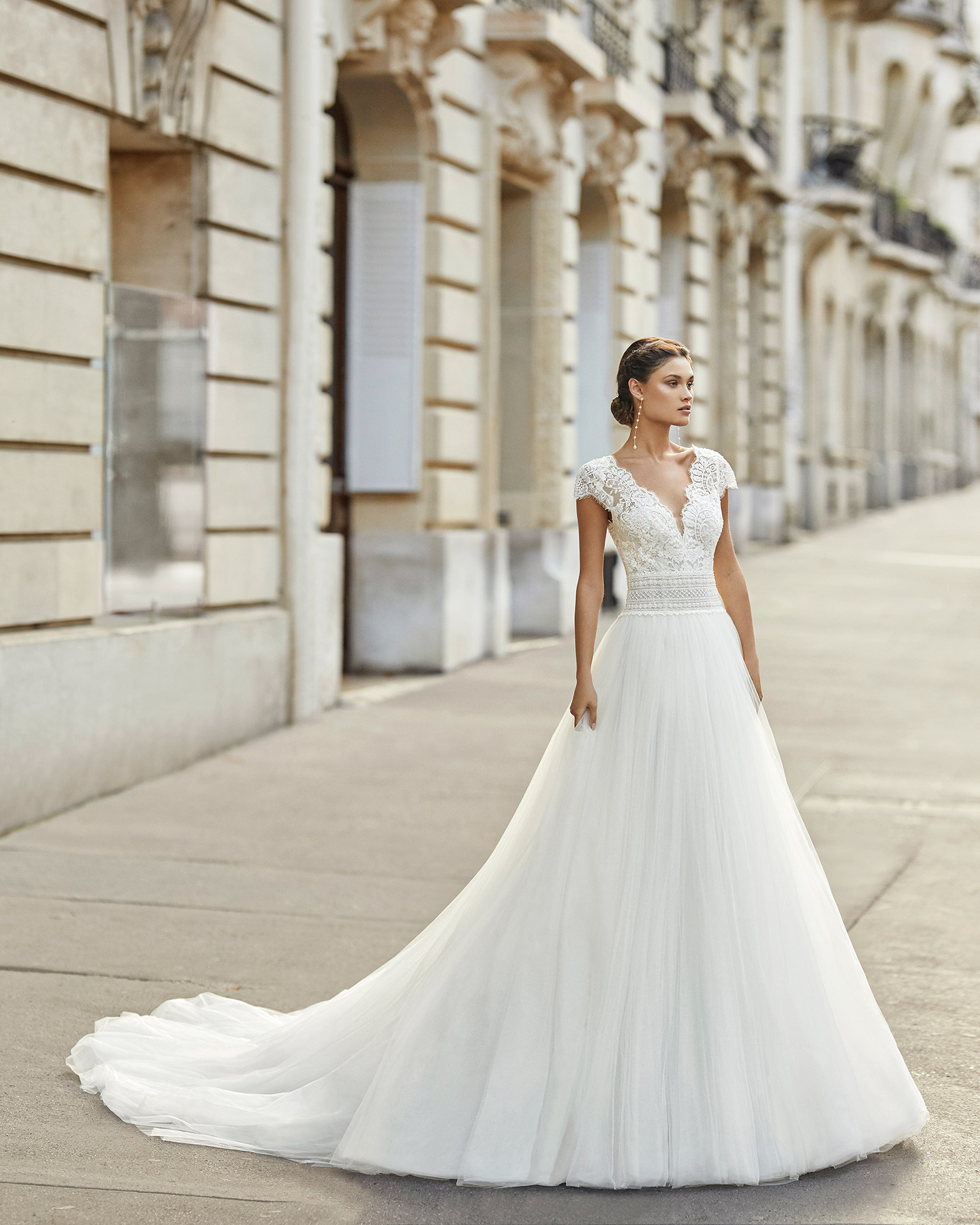 De corte princesa e um padrão de renda a acentuar a cintura, o modelo Tesalia apresenta uma saia lisa e mangas caídas, ideais para qualquer cerimónia na primavera. Exibe ainda um decote em V e costas ilusão, com renda floral, linha de botões e adorno em tule plumeti. | Rosa Clará 2021