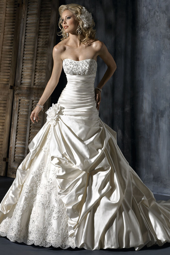 Vestido de noiva ajustado na parte superior e até à cintura, onde sai uma saia suave enfeitada com renda e uma flor na cintura.
<a href="http://www.maggiesottero.com/dress.aspx?style=A3365LU" target="_blank">Maggie Sottero Platinum 2015</a>
